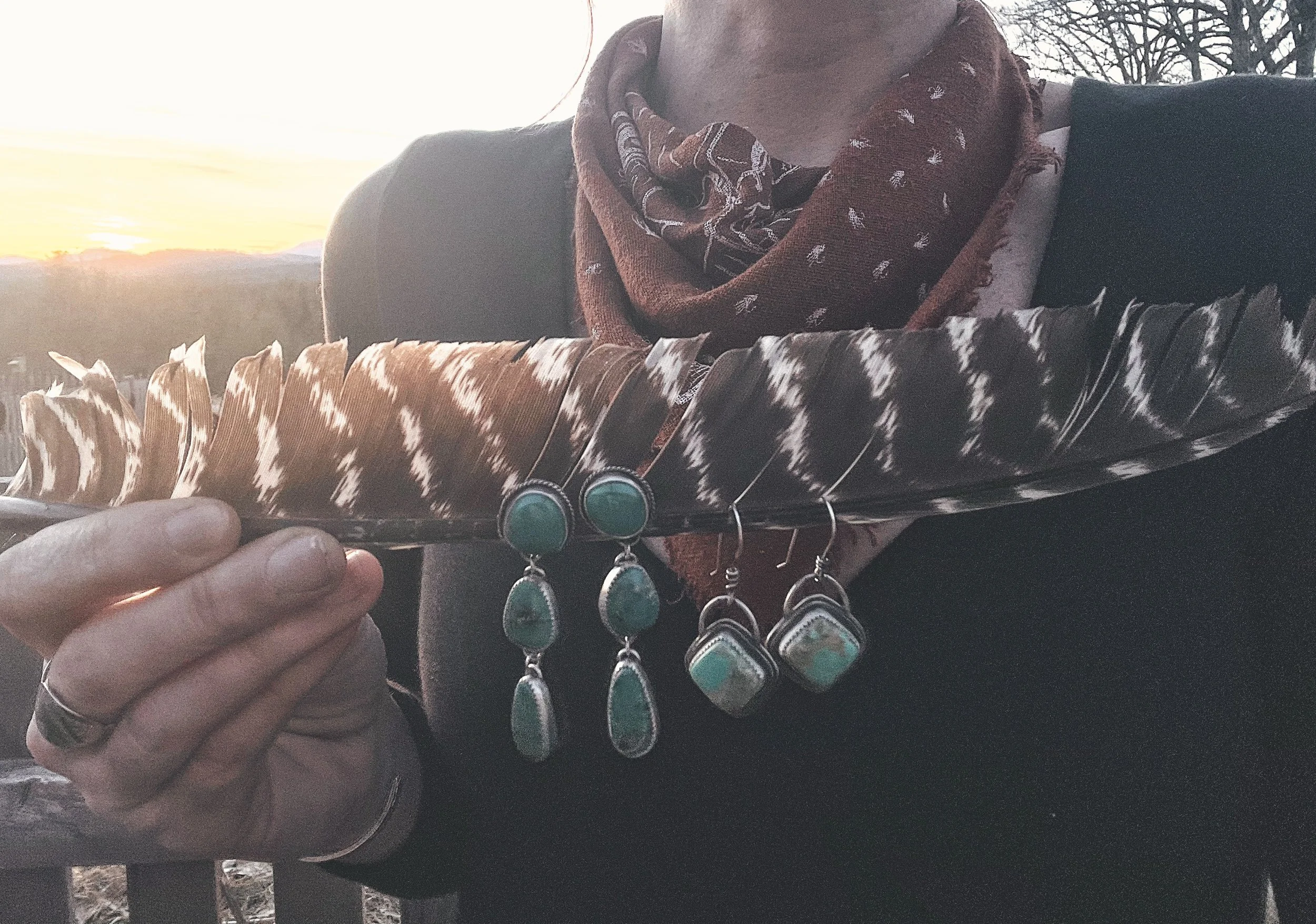 Person holding a large feather with turquoise and silver jewelry hanging from it, during sunset with trees in the background.