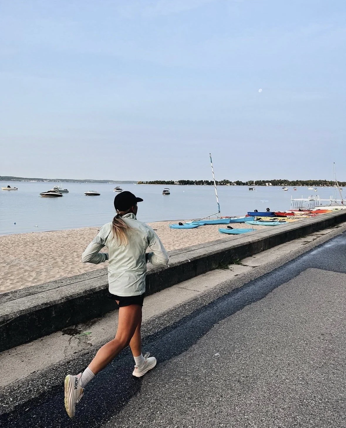 A person jogging along a waterfront promenade near a sandy beach with sailboats and motorboats in the water. The sky is clear with a visible moon.