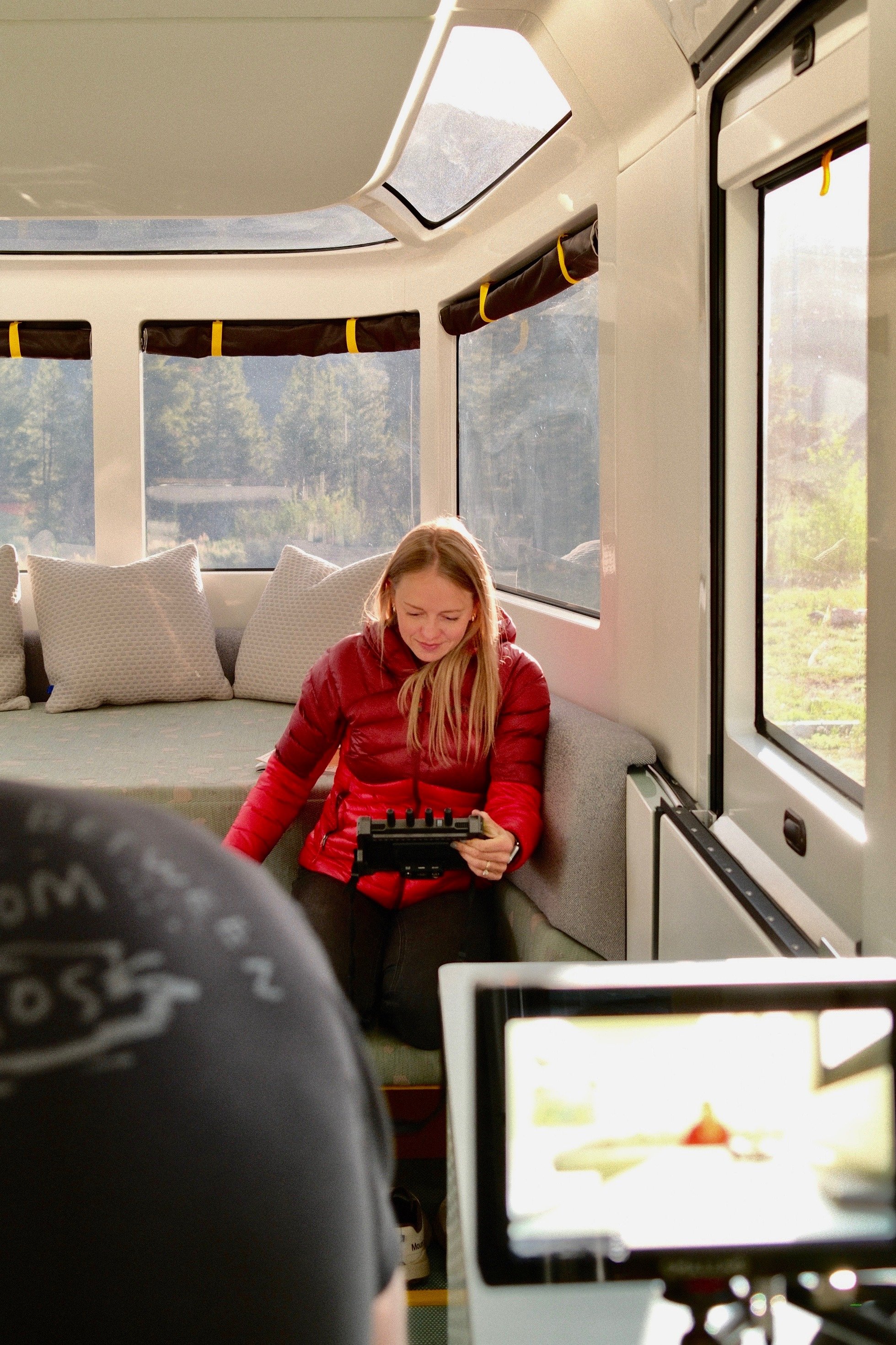 A woman sitting on the floor inside a cozy bus or camper van, surrounded by large windows showing a scenic outdoor landscape with trees and mountains, using a device in her hands.