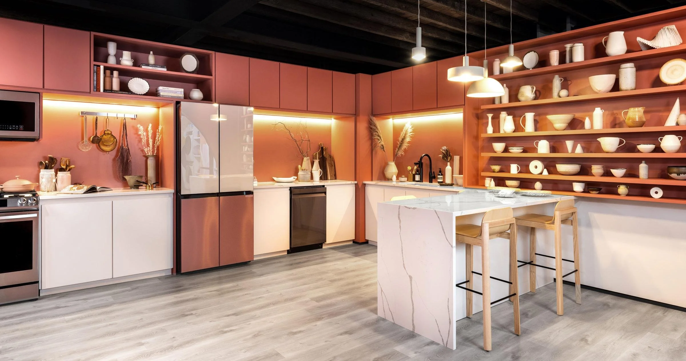 Modern kitchen with pink upper cabinets, white lower cabinets, and open wooden shelves filled with white and beige dishware, a kitchen island with a marble surface, and pendant lighting.