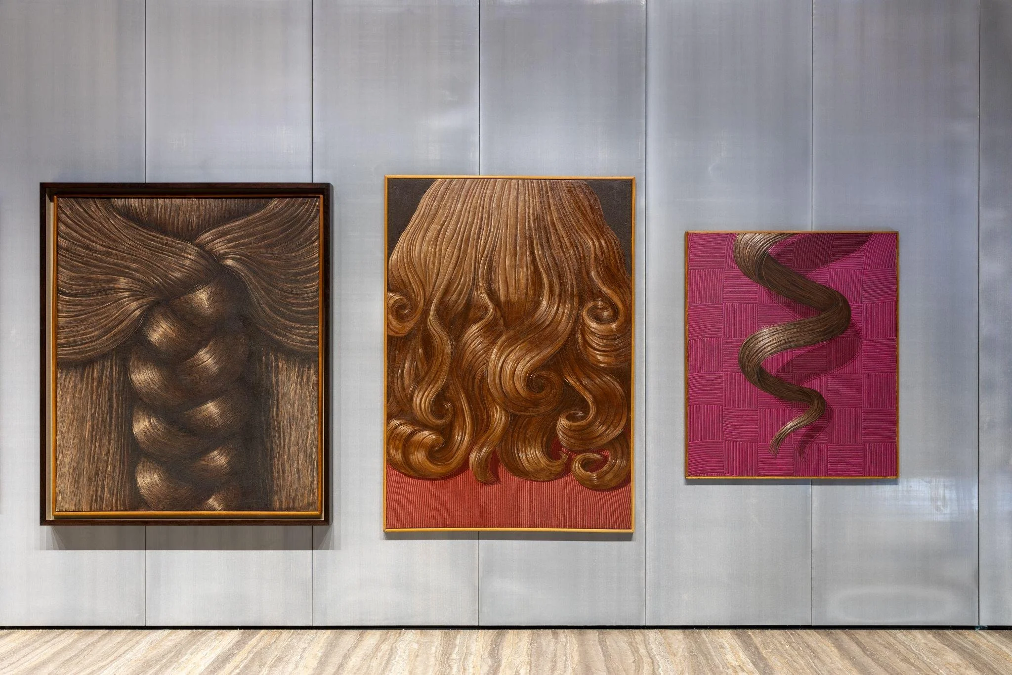 Three framed artworks featuring realistic hair designs in shades of brown and pink, displayed on a textured gray wall.