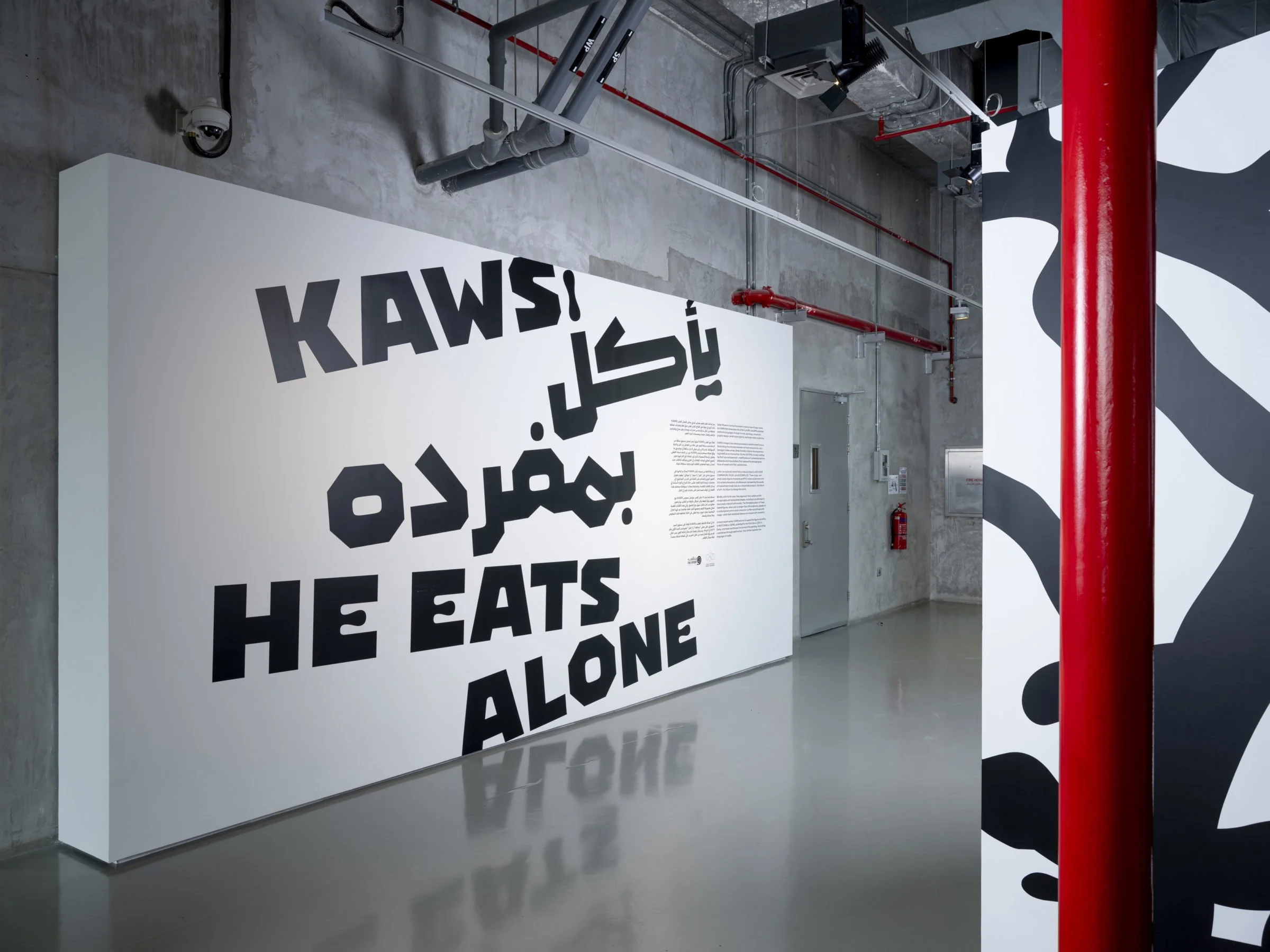 A display with large bold black letters on a white background reading, 'KAWS!'. The display is in an industrial space with exposed concrete walls and ceiling, pipes, and security cameras.