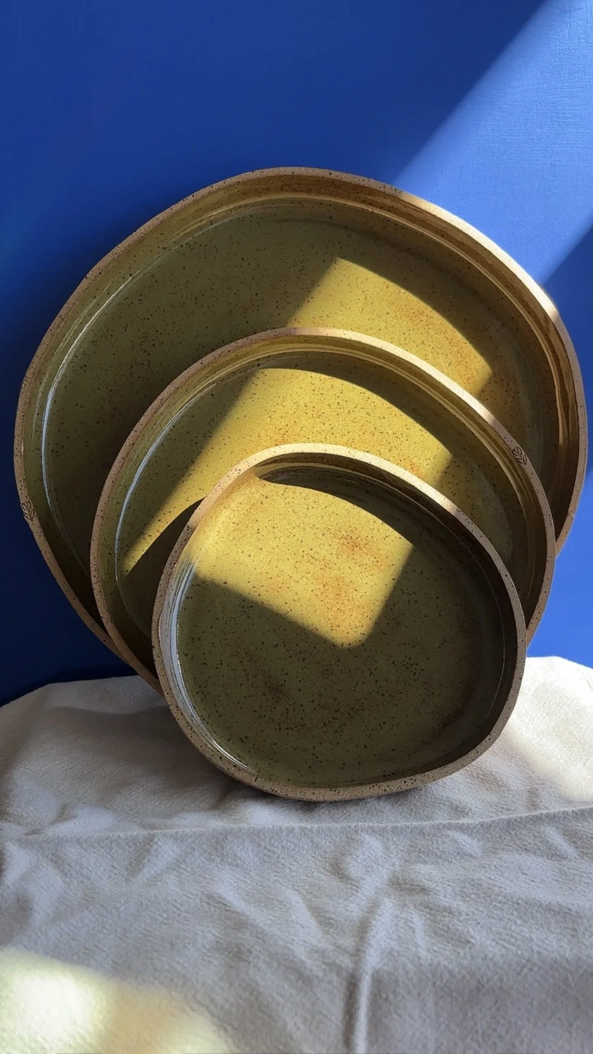 Three round shallow bowls with a speckled yellow-green glaze stacked inside each other, placed on a white cloth with blue background and sunlight casting shadows.