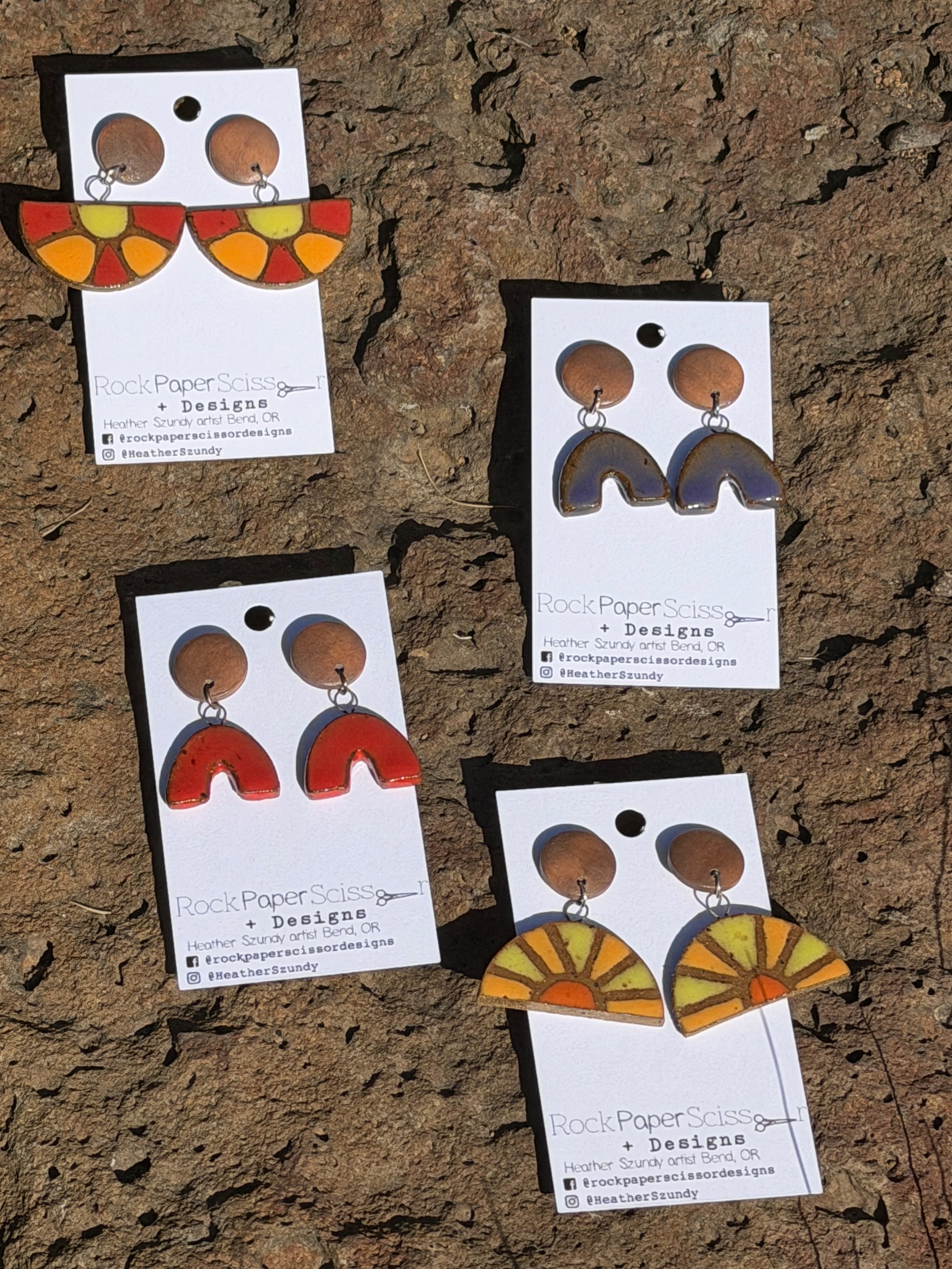 Colorful earrings displayed on white cards with business information for Rock Paper Scissors + Designs, featuring an artist from Bend, Oregon. The earrings are designed to resemble semi-circular, geometric patterns with various colors and styles.