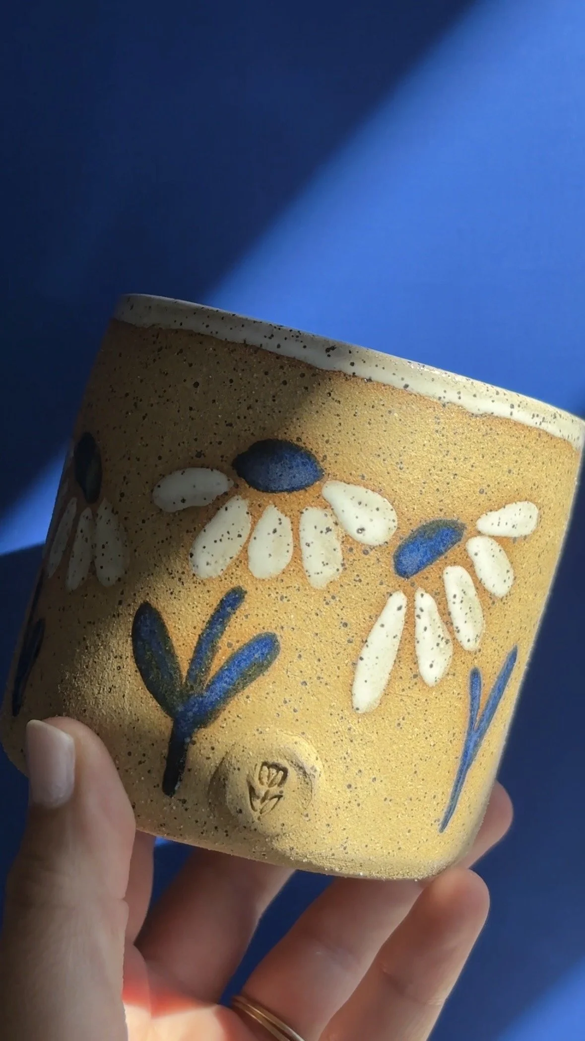 Close-up of a ceramic cup featuring a floral pattern with white and dark blue petals and leaves on a yellow background, held in a person's hand against a blue background.