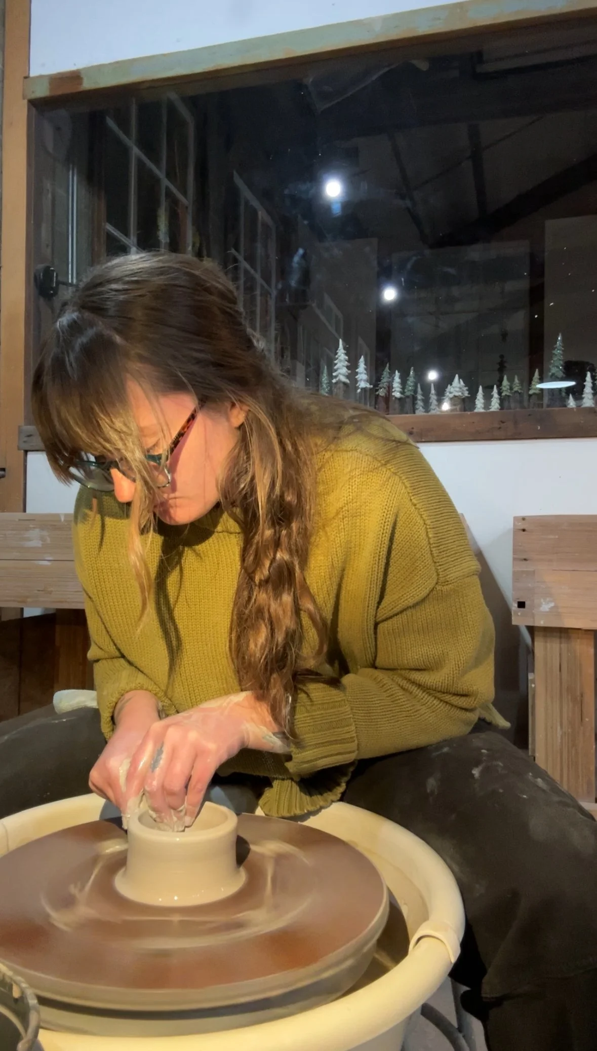 A woman with glasses and long hair, wearing a mustard yellow sweater, is working on pottery at a pottery wheel in a studio. Through the window behind her, a nighttime scene features a full moon and snowy pine trees.