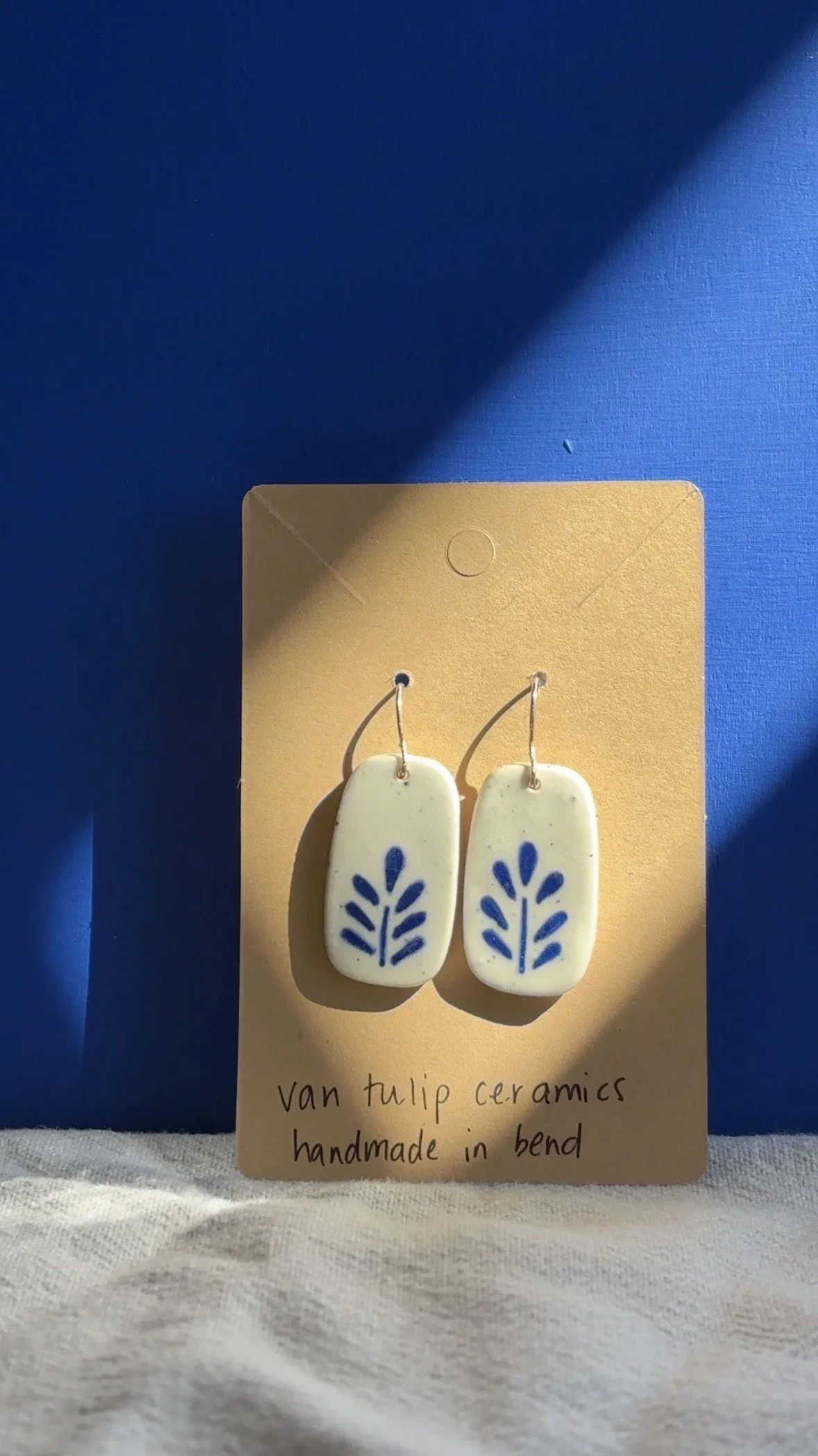Pair of handmade ceramic earrings with blue tulip floral pattern hanging on beige card, with a blue and beige background.