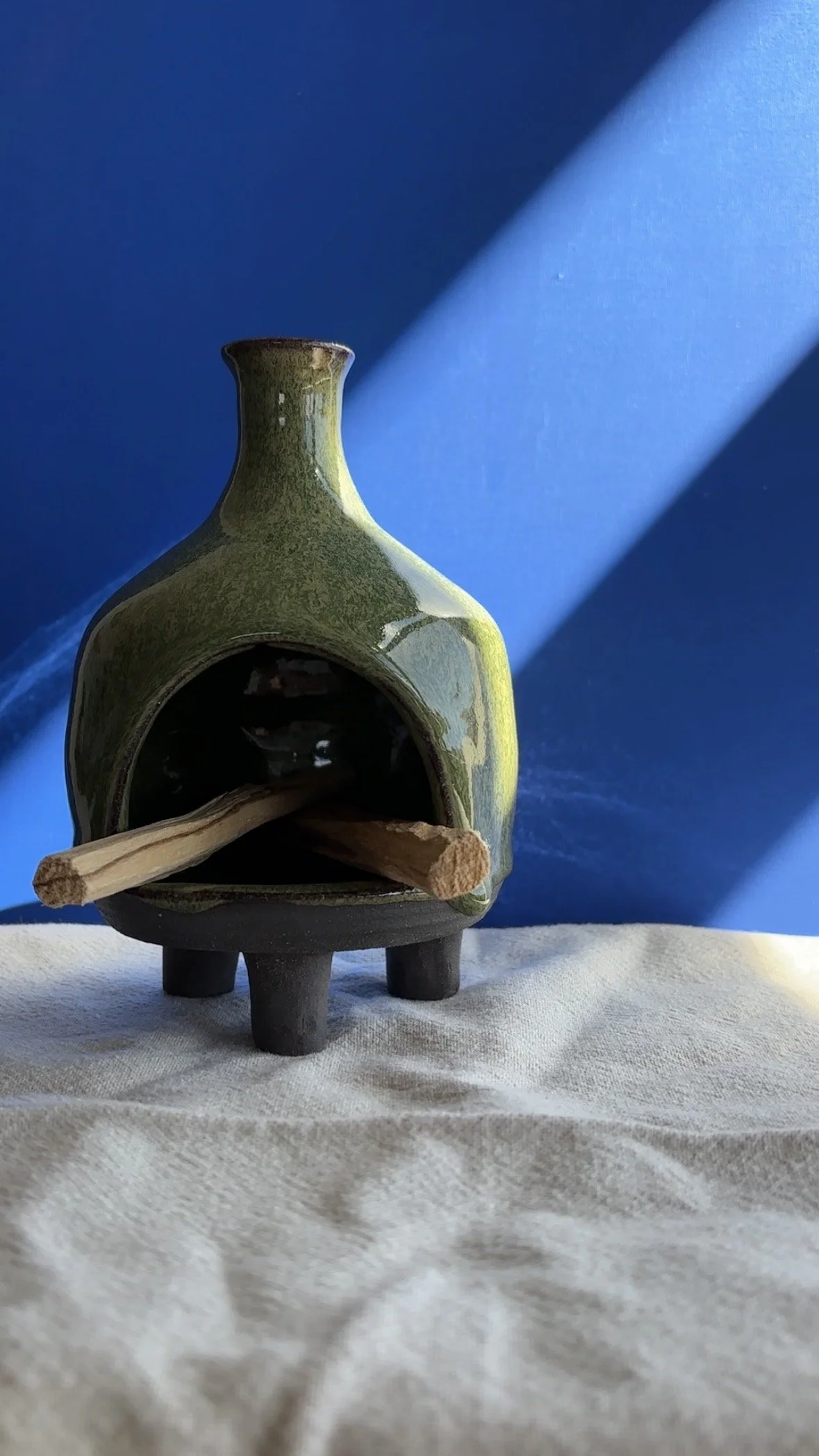 A ceramic sculpture of a small animal with a green glaze, featuring an open mouth holding a wooden stick, standing on four short legs against a blue background with sunlight casting shadows.
