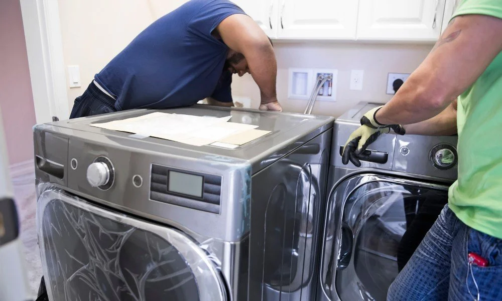 Two workers installing a washing machine in a laundry room with white cabinets and outlets on the wall.