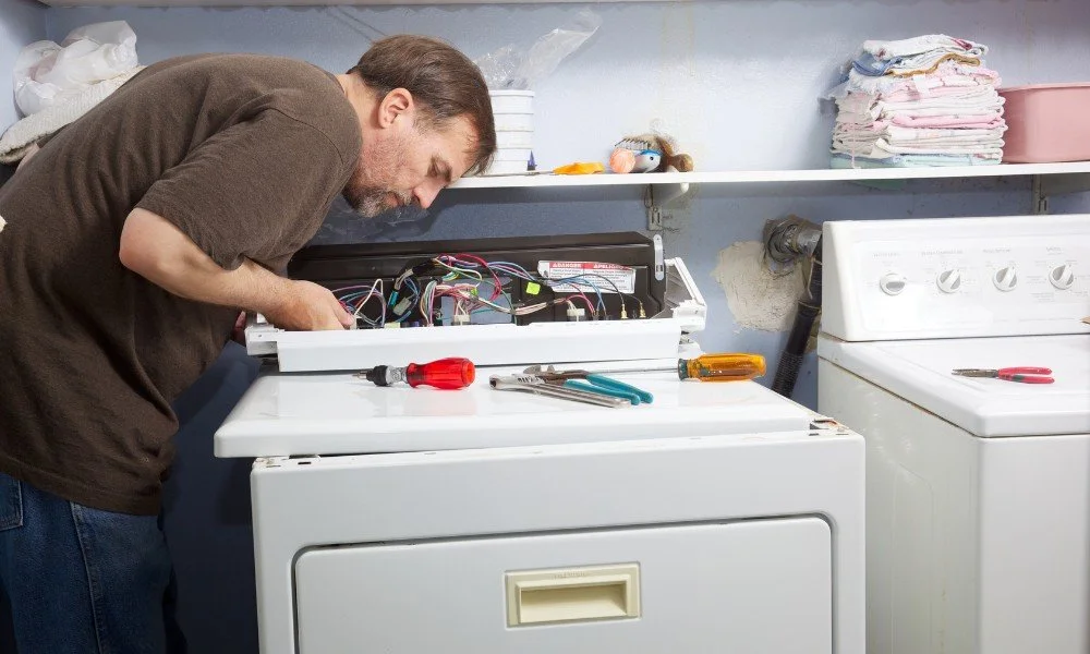 Man repairing a washing machine in laundry room with tools on top of the machine.