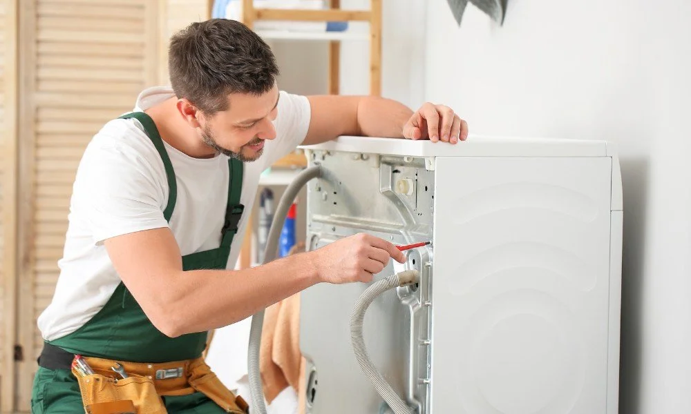 A repairman fixing a washing machine with a screwdriver in a laundry room.