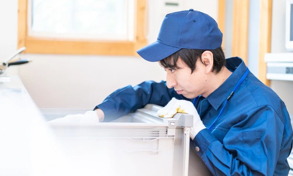 A male worker in a blue uniform and cap cleaning a dishwasher with a cloth in an office or kitchen setting.