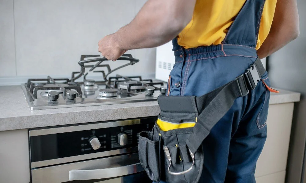 A worker in a yellow shirt and blue overalls with a tool belt is working on a gas stove in a kitchen.