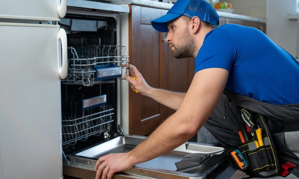 A technician in a blue shirt and cap inspecting an open dishwasher with tools attached to his tool belt.