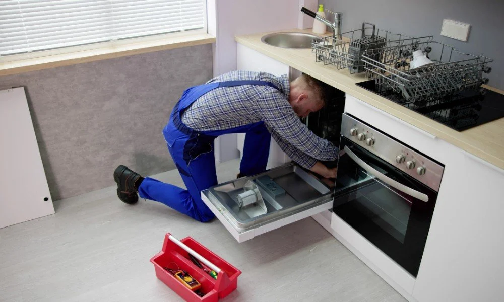 A man in blue overalls kneeling in front of an open kitchen oven, repairing or inspecting the appliance. Kitchen has a countertop with dish racks, a sink and some tools in a red toolbox on the floor.