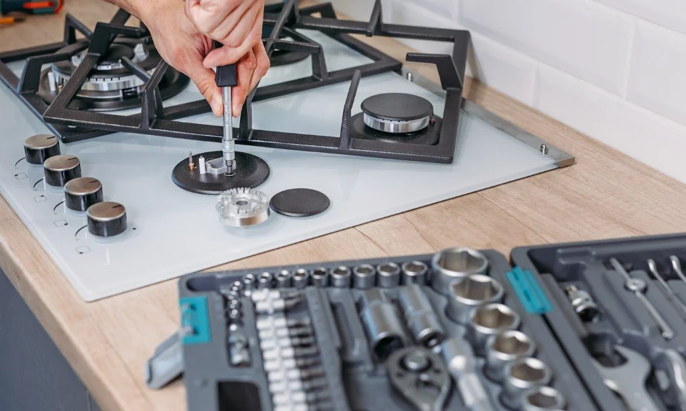 A person using a screwdriver to assemble or repair a black gas stove, with a toolbox containing various socket wrenches and tools nearby.