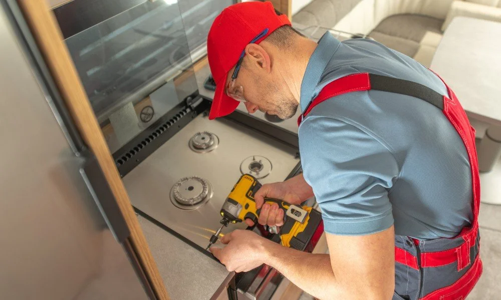 A handyman wearing a red cap, glasses, and blue work shirt with red suspenders is installing or repairing a kitchen stove using a yellow cordless drill.