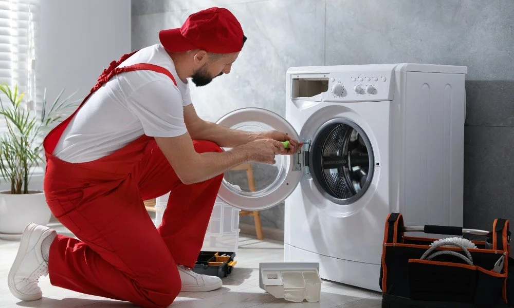 A man in red overalls and a red cap repairing a washing machine, kneeling on the floor with tools nearby.