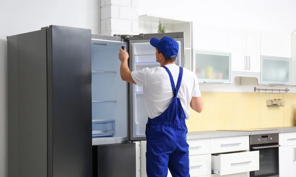 A man wearing a blue cap and overalls cleaning an open refrigerator in a modern kitchen