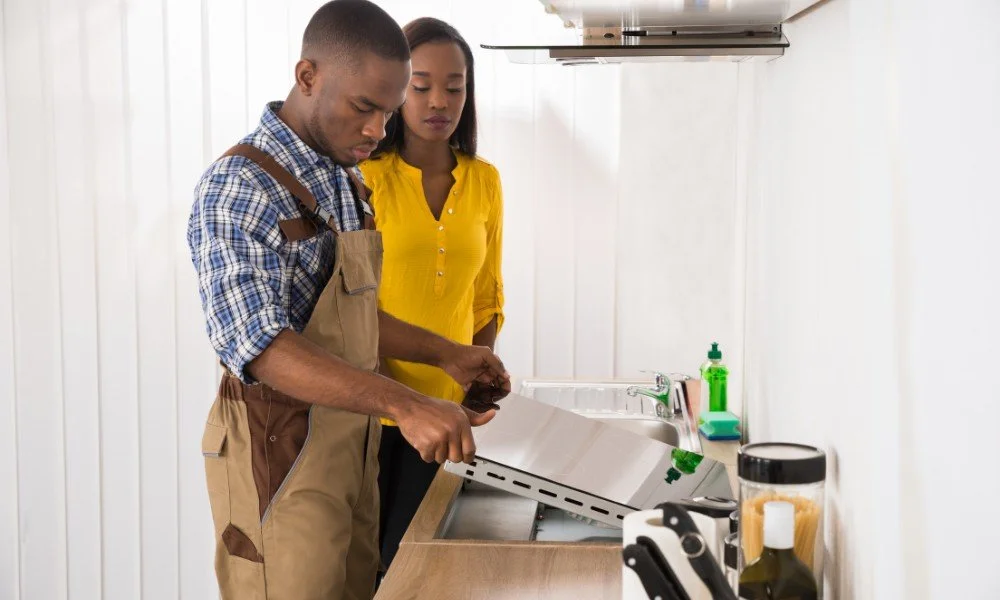 A man and woman in a kitchen, with the man breaking a chocolate bar. The woman is watching him. The man is wearing an apron over a checkered shirt, and the woman is wearing a yellow blouse.