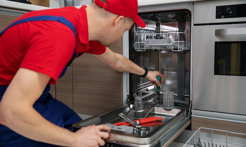 A technician repairing a dishwasher. The technician is wearing a red cap and red shirt, using a screwdriver inside the opened dishwasher, which has various tools on the kitchen counter.