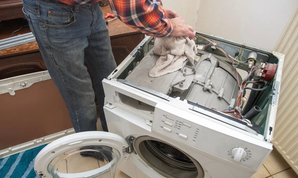 Person repairing a washing machine with the top panel removed, using a cloth to clean or check inside.