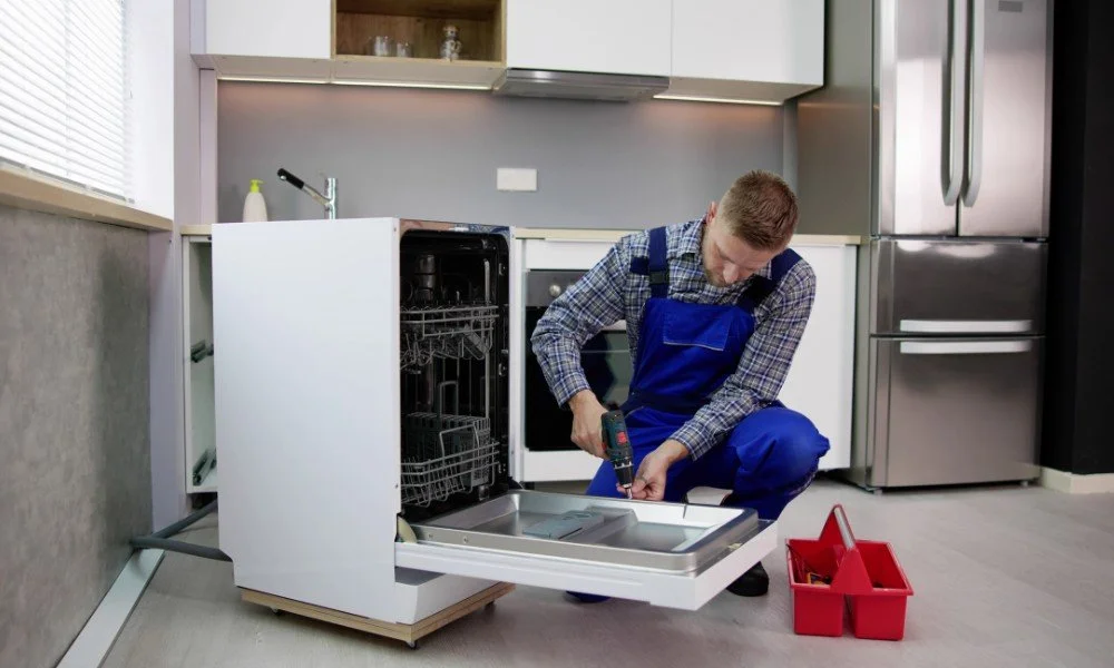 A man repairing a dishwasher in a modern kitchen with stainless steel appliances, using a screwdriver with a red toolbox nearby.