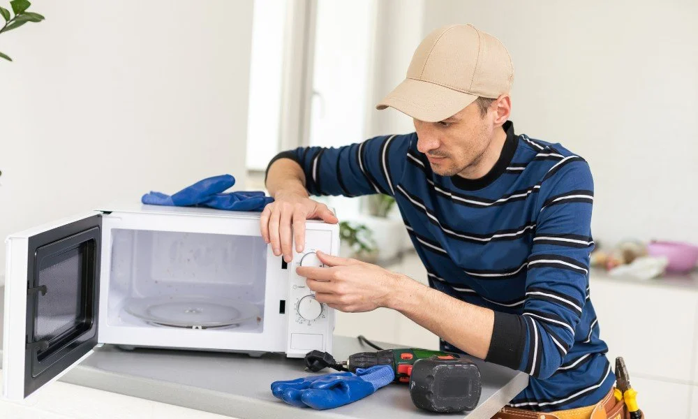 A man in a striped shirt and tan cap working on a microwave with tools and gloves on a white countertop.