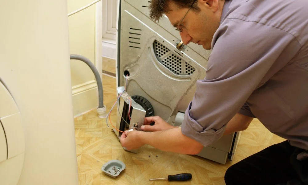 A man repairing the back of a washing machine on a wooden floor.