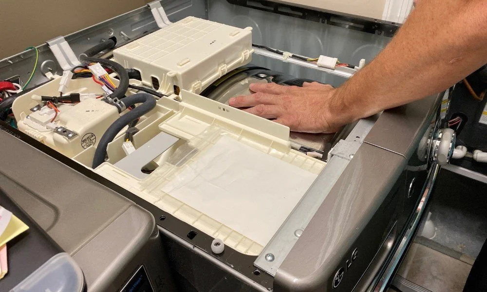 Technician working on the internal components of a washing machine with various electrical parts and wiring visible.