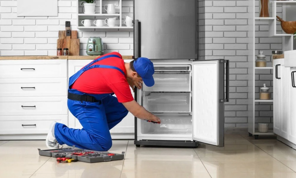 A maintenance technician kneeling in front of an open refrigerator in a modern kitchen, performing repairs with tools.
