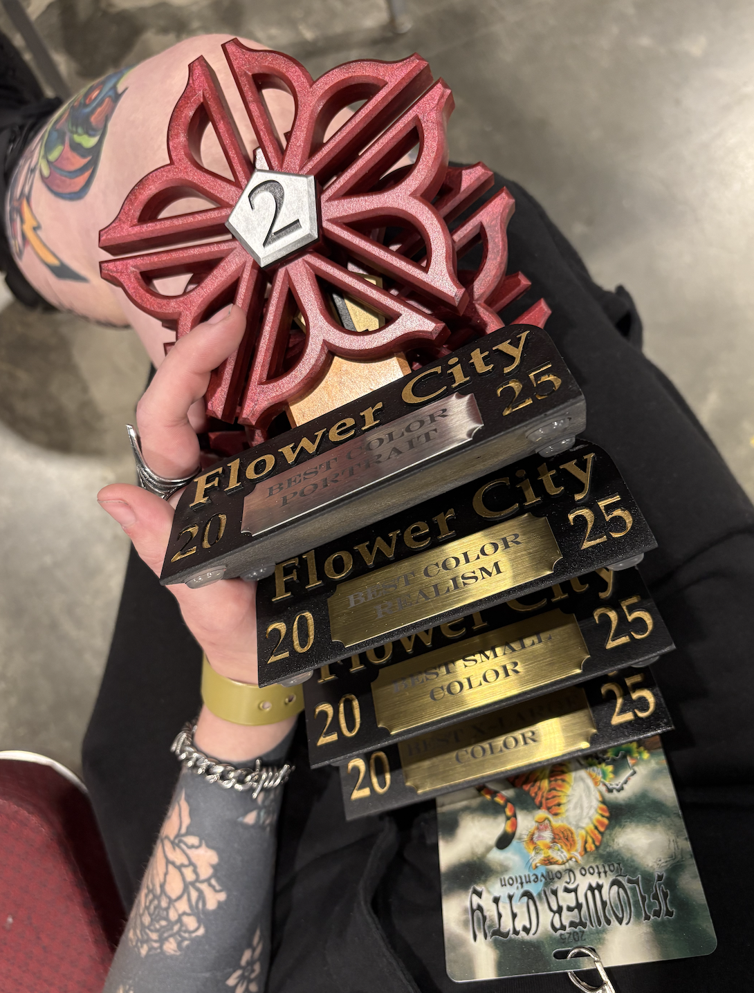 Person holding multiple awards and trophies, including a red flower-shaped award with a silver hexagon numbered 2, and several black and gold awards labeled 'Flower City' and 'Best Color REALISM'. The person has tattoos and is wearing a yellow watch and silver bracelet.
