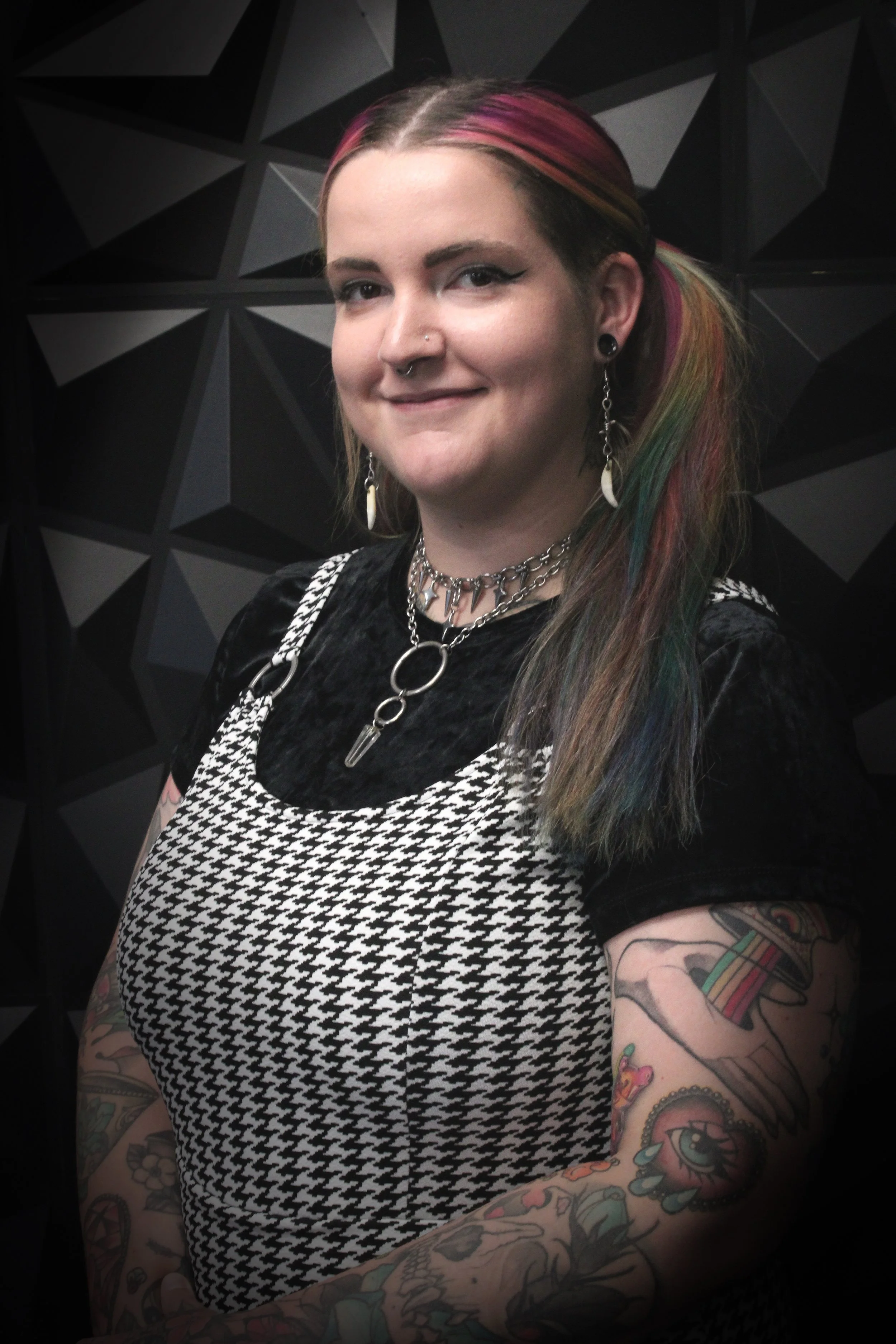 A woman with rainbow-colored hair, tattoos on her arms, and multiple piercings, standing against a black geometric background.