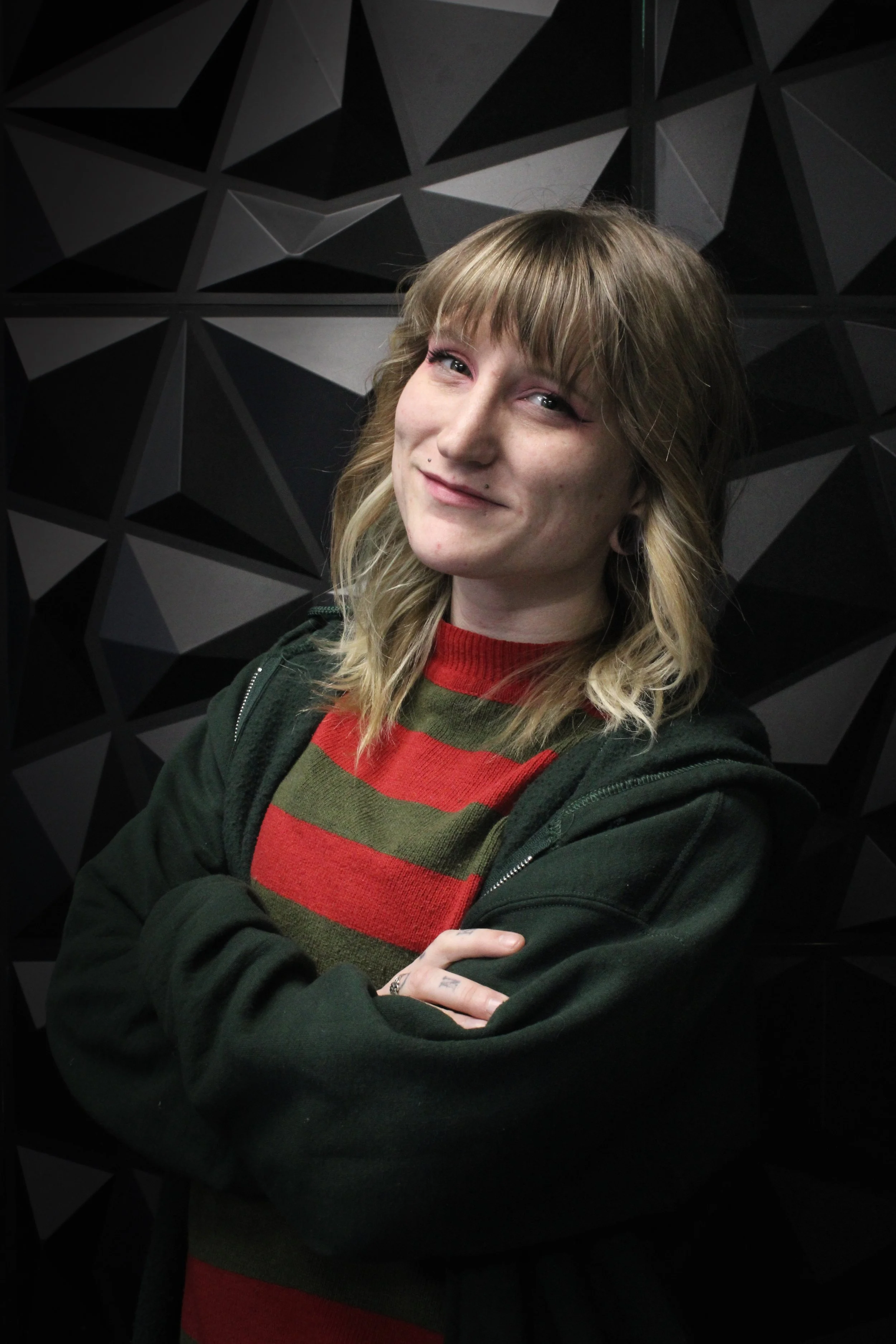 A young woman with shoulder-length blonde hair, wearing a red and green striped sweater and a black hoodie, stands with arms crossed in front of a black, geometric-patterned wall.