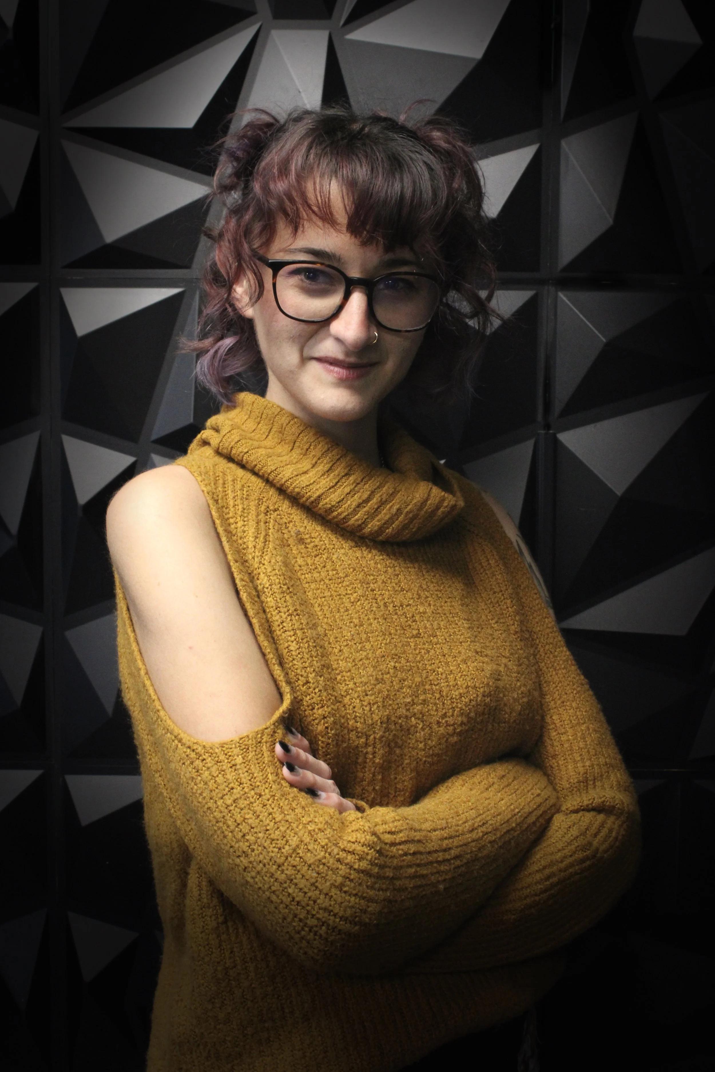 A woman with glasses and curly, purple-tinted hair, wearing a mustard yellow, cold-shoulder sweater with her arms crossed, standing against a black geometric patterned background.