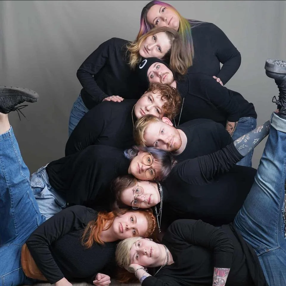 A group of ten young people stacked in a pyramid formation against a gray background, with colorful hair, glasses, tattoos, and casual clothing.