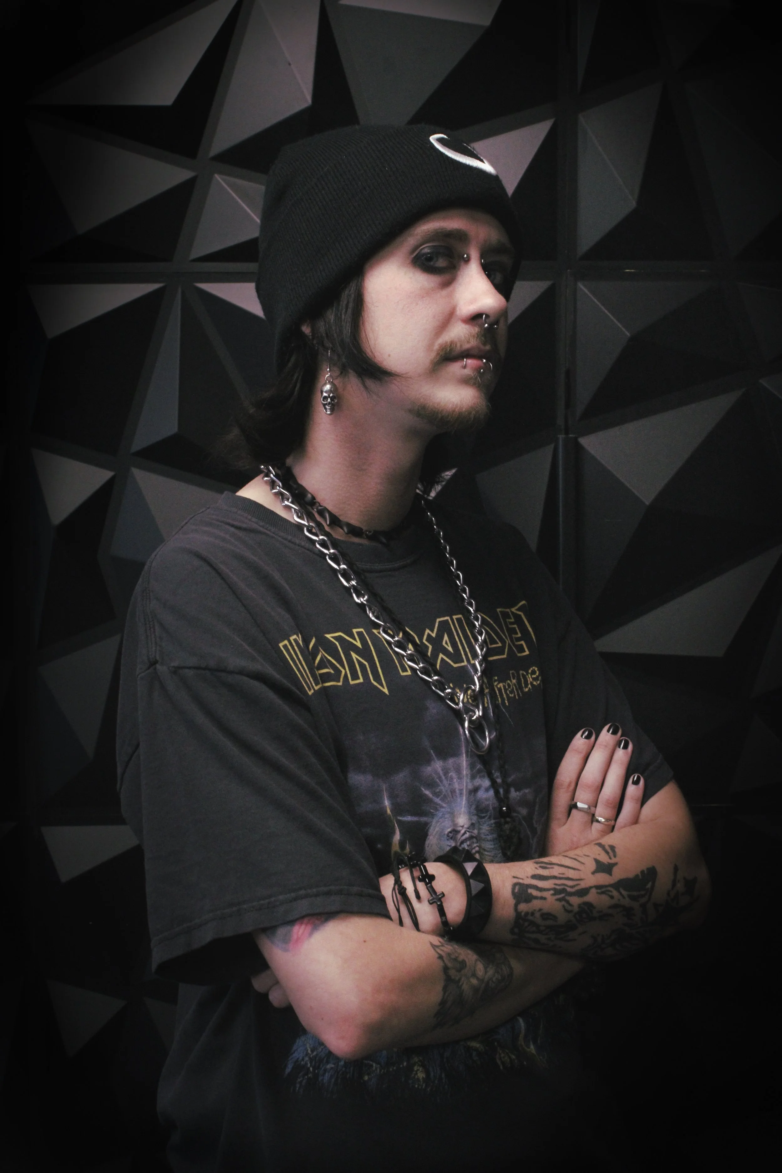 A person with long dark hair wearing a black beanie, dark makeup, and tattoos on their arms, dressed in a black Metallica T-shirt, standing with arms crossed against a black geometric wall.