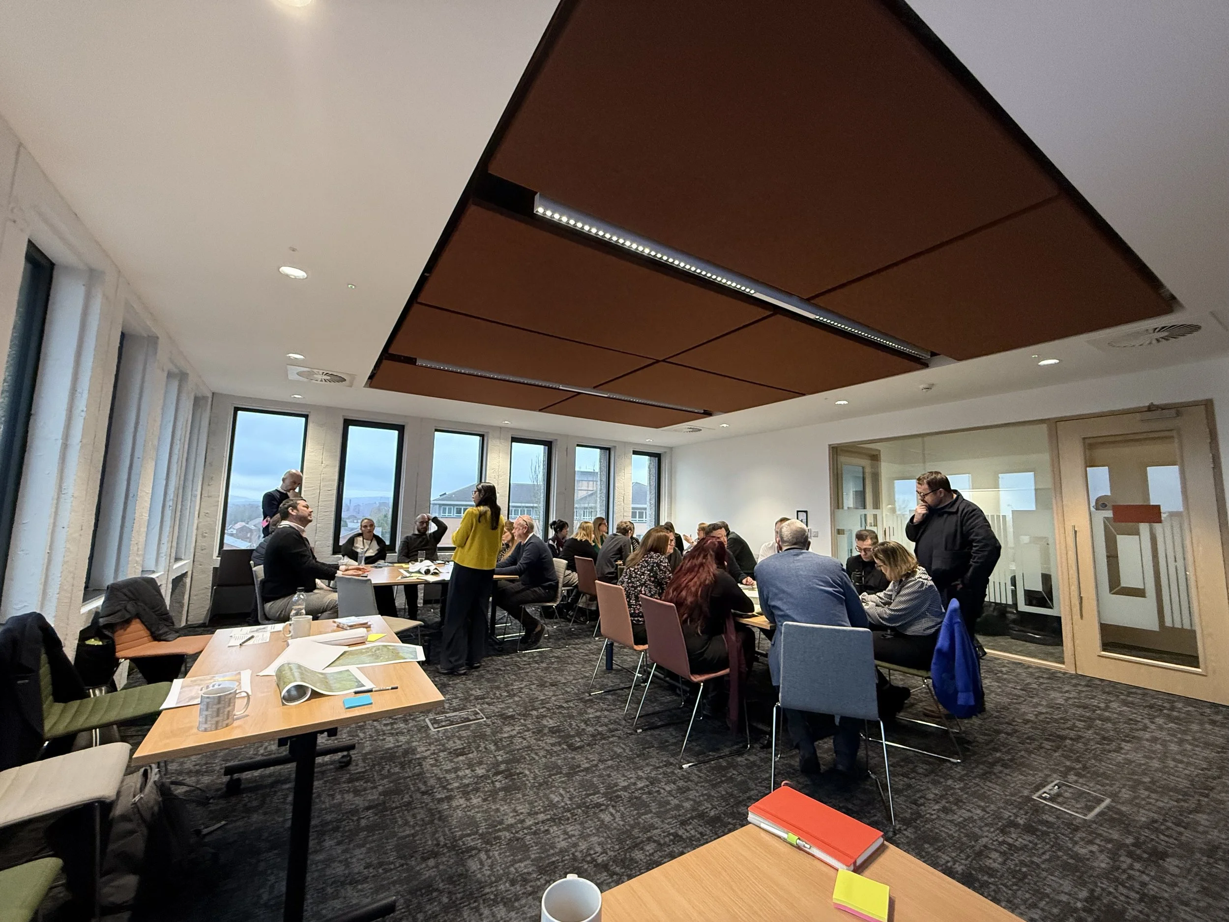 A group of people are gathered in a conference room with large windows, sitting around tables, engaged in discussion and listening to a presenter. Some are standing and others are seated, with books, papers, and cups on the tables. The ceiling has brown panels, and the room has a modern, professional appearance.