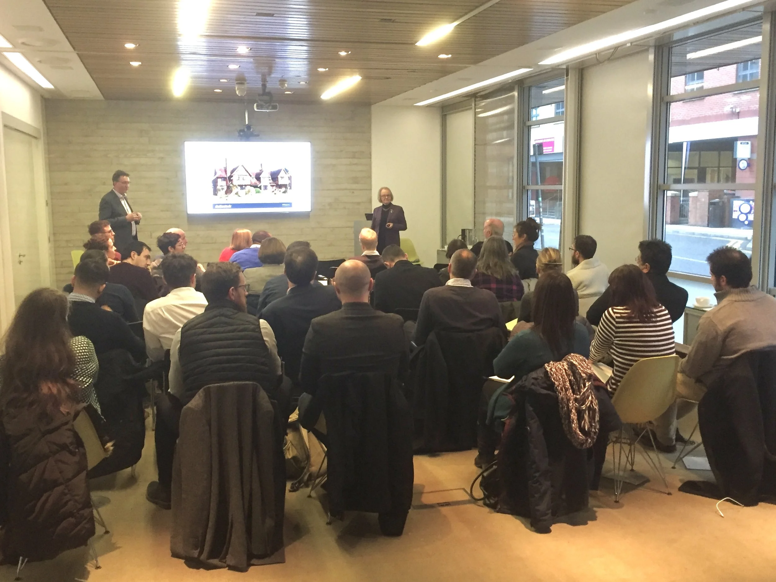 People attending a presentation in a conference room with large windows, seated and listening to a speaker in front of a screen displaying a slide.