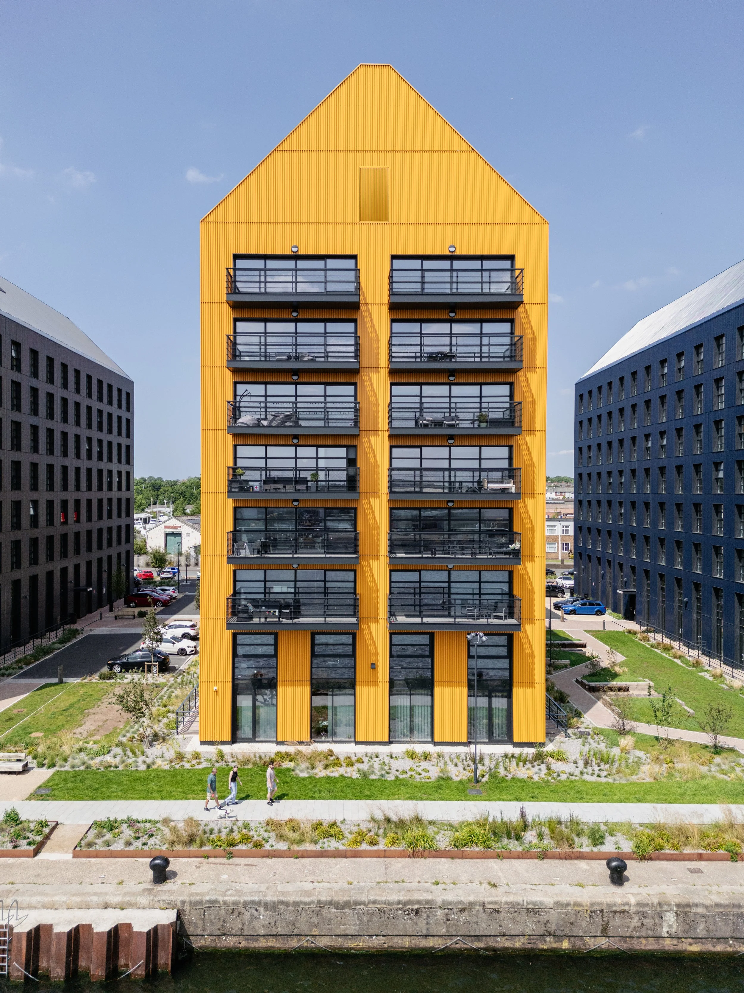 Modern yellow and black apartment buildings with balconies, situated near a river with pedestrians walking on a pathway.