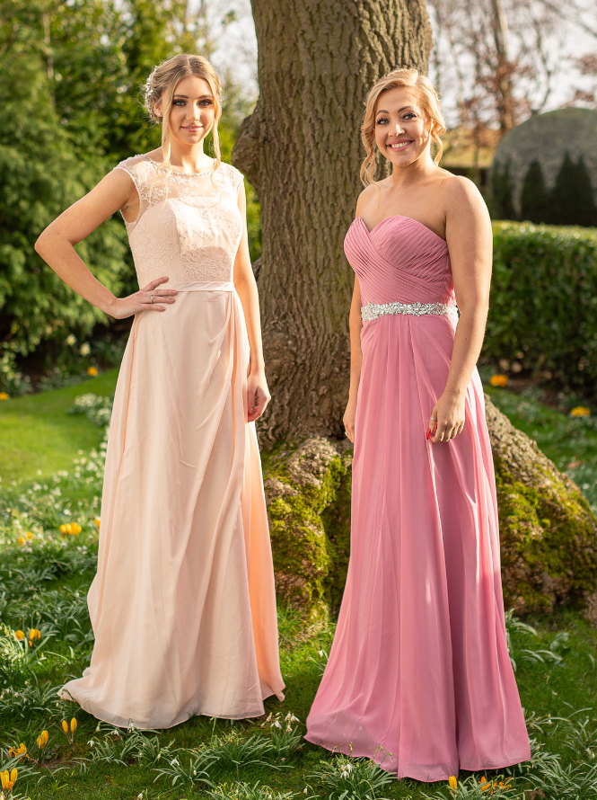 Two women wearing long, elegant dresses standing outdoors near a tree in a garden. One woman in a light peach dress, the other in a pink dress with a silver belt.