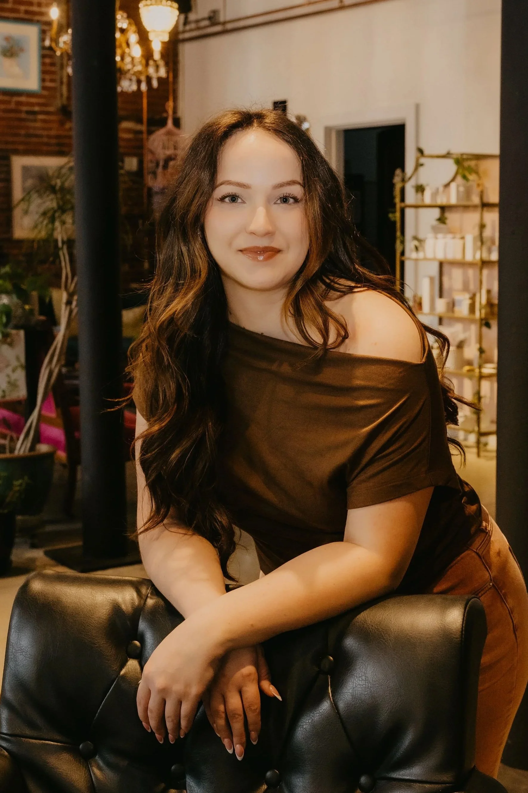 A young woman with long brown hair and light skin, wearing a black top and jewelry, smiling softly at the camera.