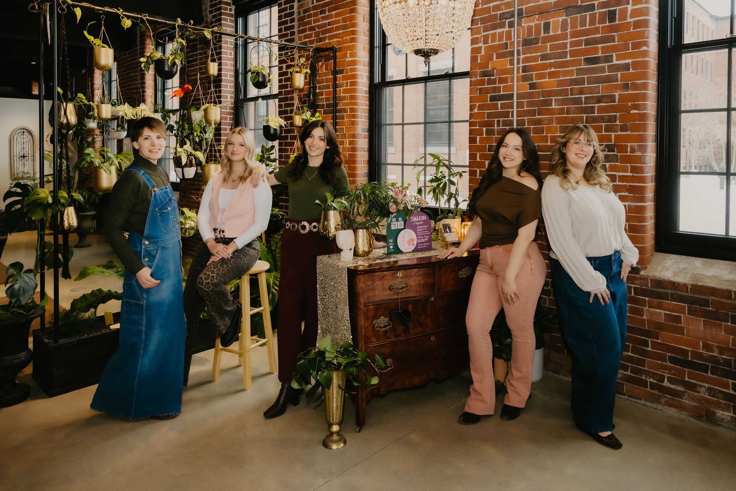 Five stylists at The Hair Garden boutique hair salon in Biddeford, Maine, posing together in a lush, plant-filled salon interior with exposed brick walls, large windows, and warm eclectic décor.