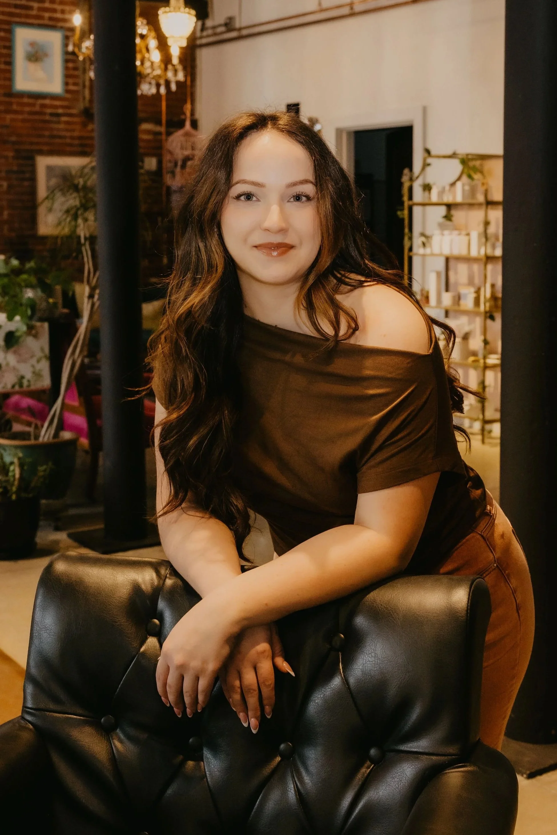 A young woman with long brown hair and light skin, wearing a black top and jewelry, smiling softly at the camera.
