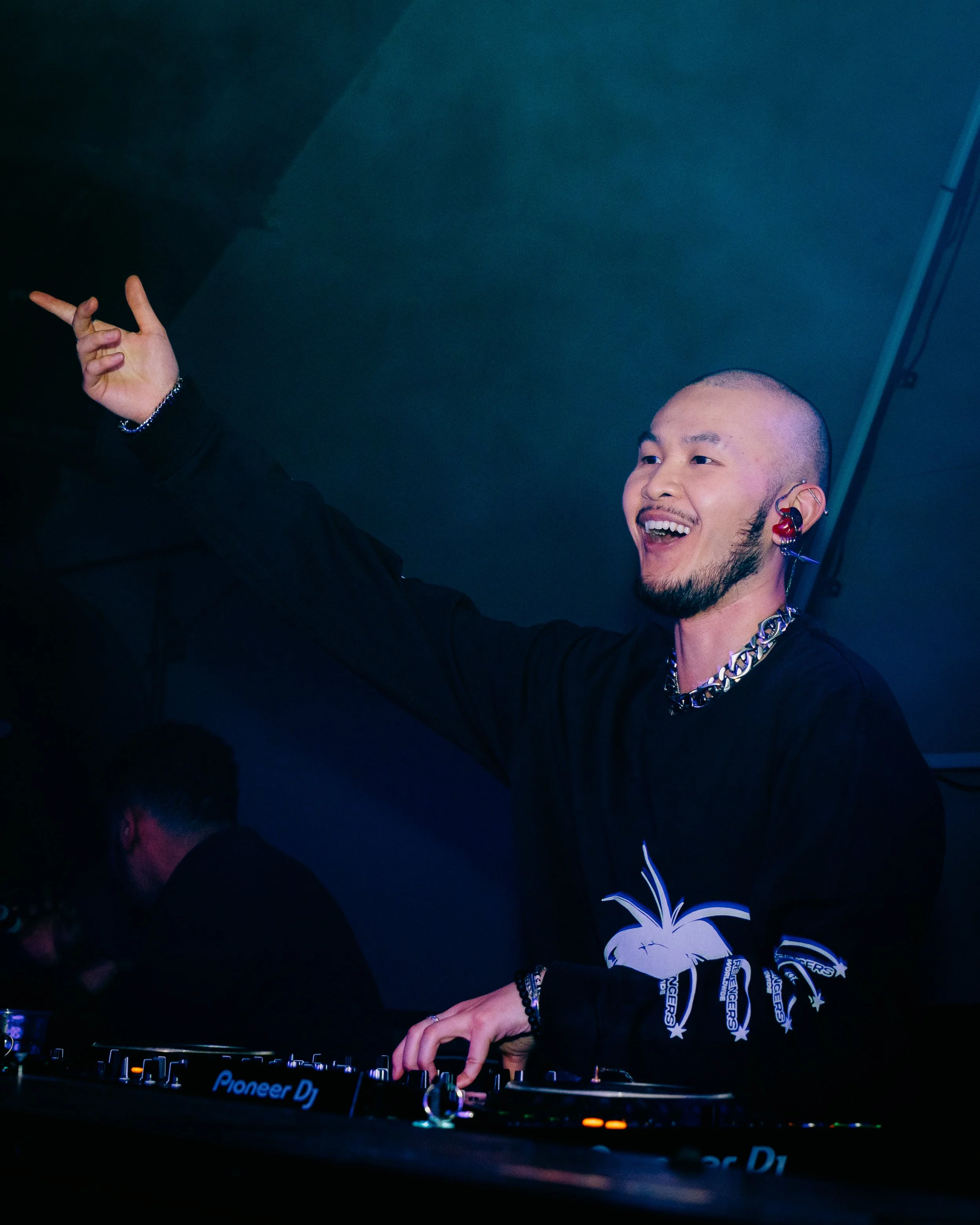 A DJ smiling and pointing with one hand while playing music at a club, wearing a black shirt with a graphic on the sleeve and jewelry.