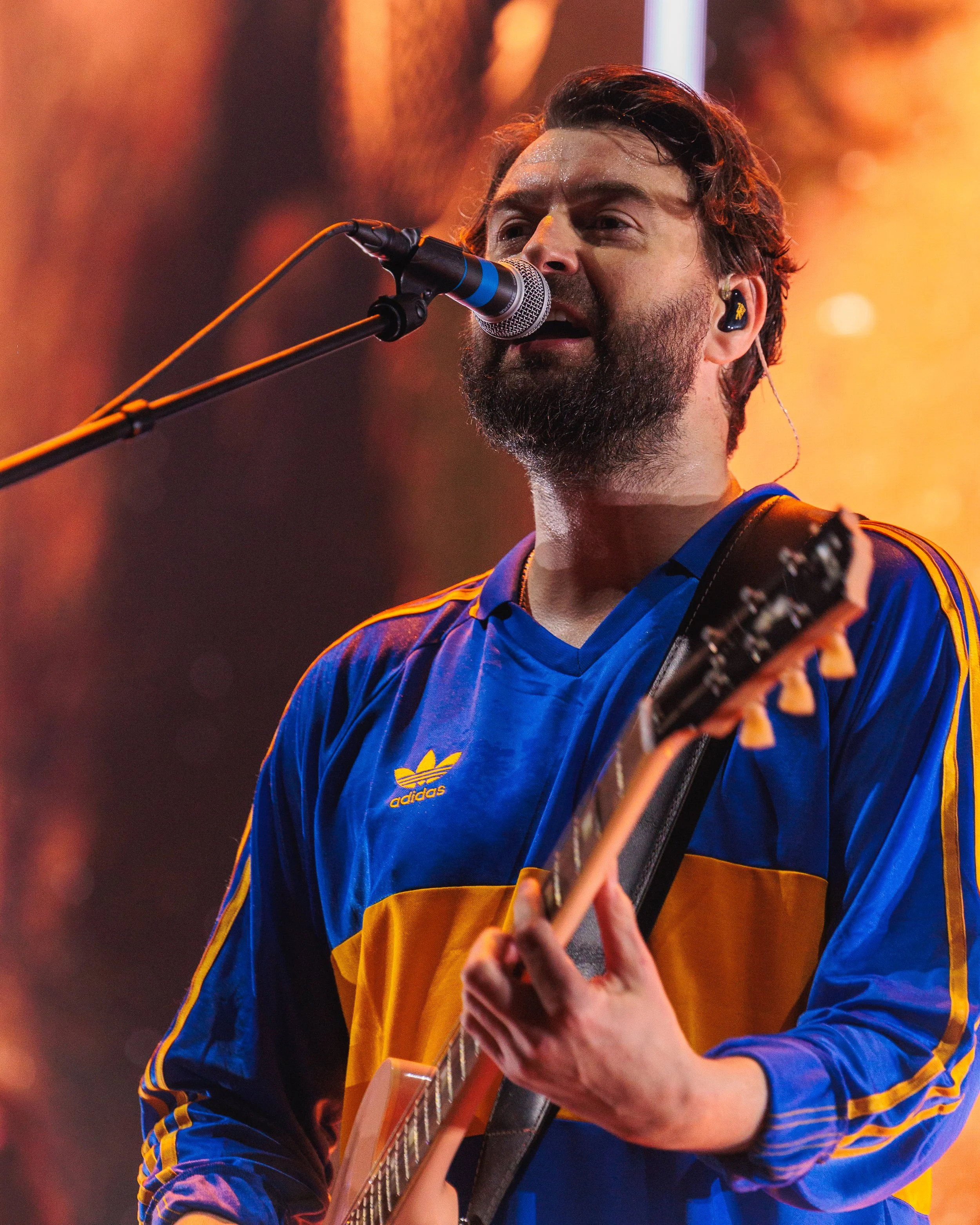 A man with a beard singing into a microphone while playing an acoustic guitar, wearing a blue and yellow Adidas jacket.