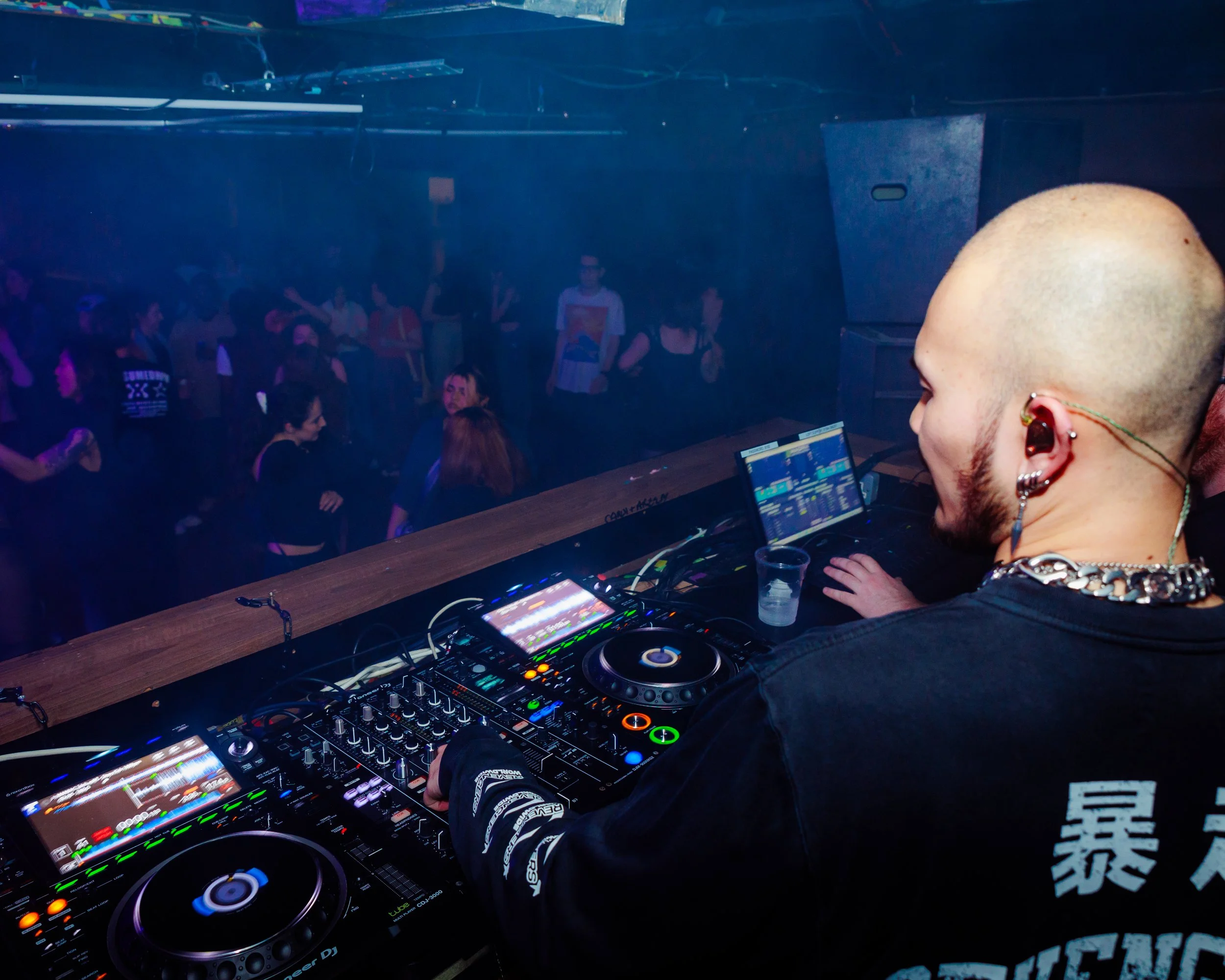 A DJ with a shaved head and earrings is performing at a nightclub. He's using DJ equipment and a laptop, with a crowd of people dancing in the background.