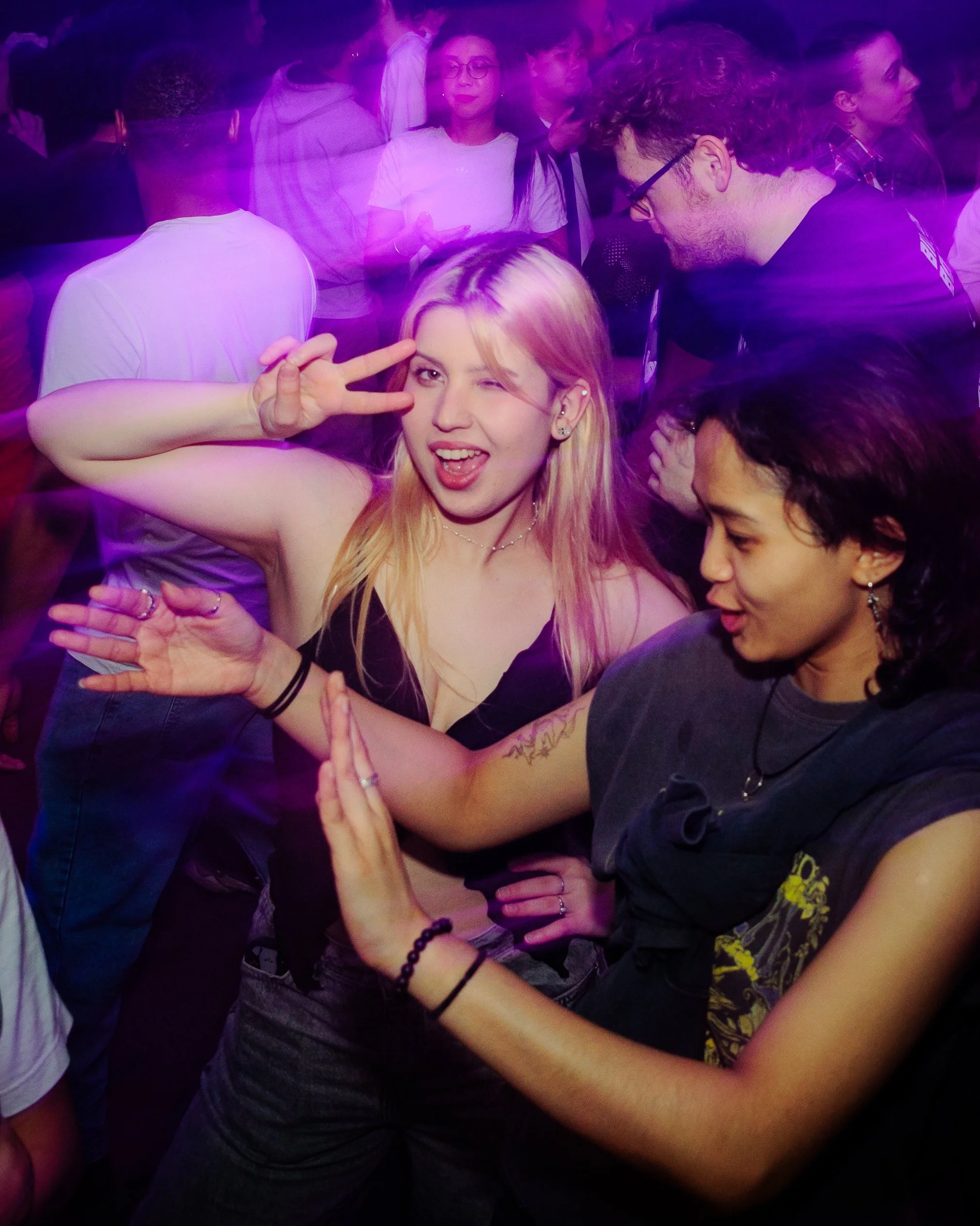 A group of young people dancing at a party with purple lighting. A blonde girl in the center is making a peace sign with her fingers and winking, while the person next to her is giving a high five.
