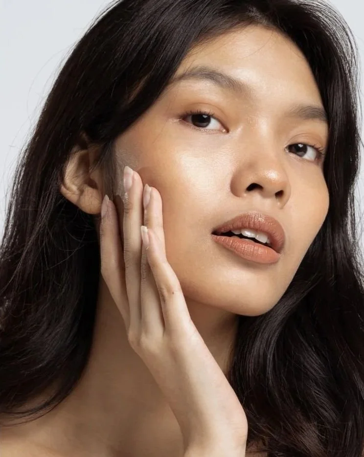 Close-up of an Asian woman with clear, smooth skin touching her cheek, looking at the amazing results of a Dermabrasion facial at Flirt Custom Lash Studio Toronto.