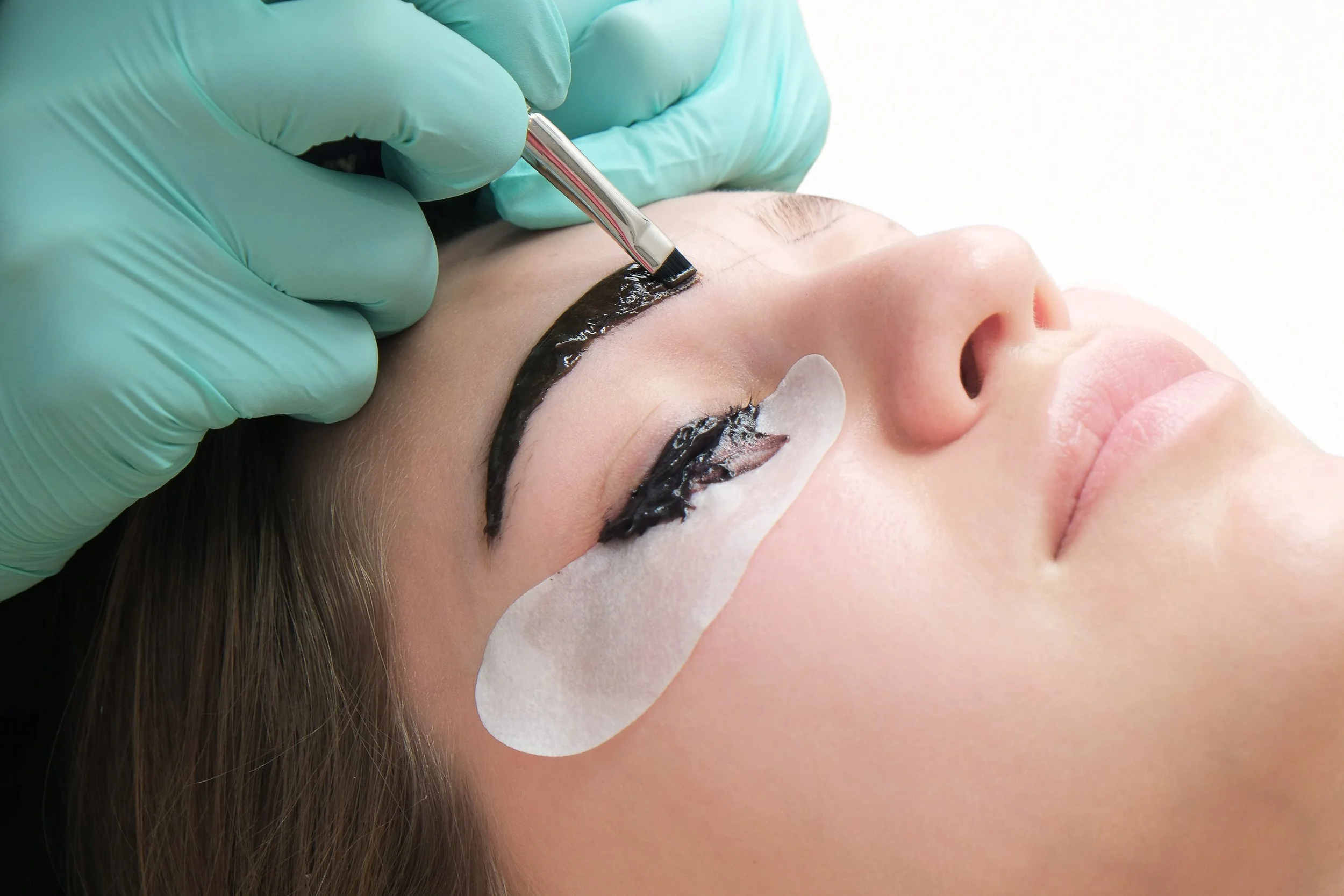 A technician in teal gloves applies brow product as a client receives a brow lamination and lash lift at Flirt Custom Lash Studio. Under-eye pads protect the skin during the treatment.